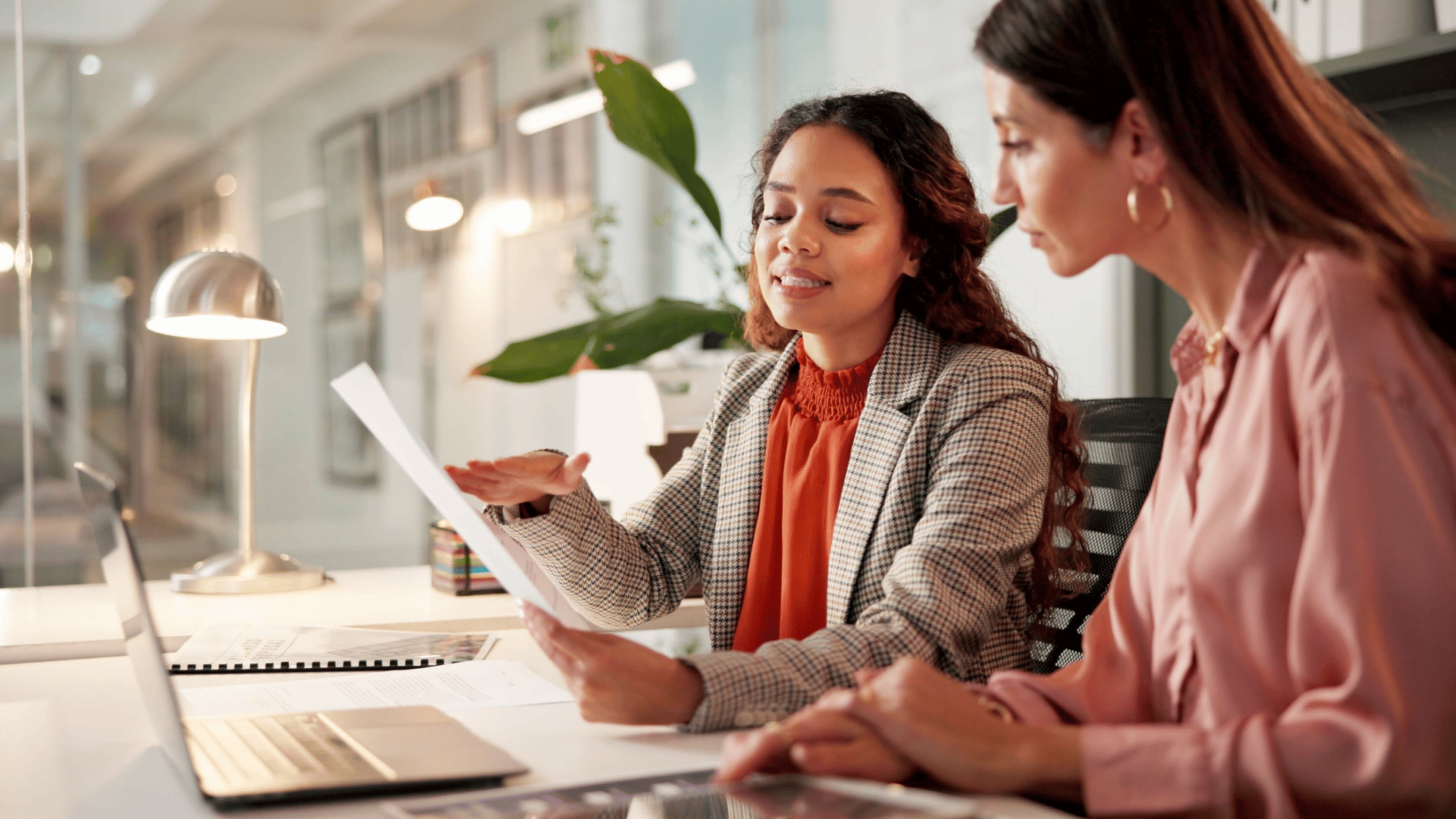 Financial Education Two woman dressed in business casual attire site a table reviewing a document in front of a laptop.